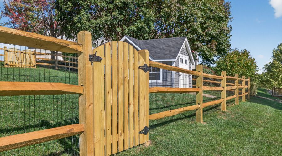 Wooden fence with gate in a grassy yard beside a gray house under trees
