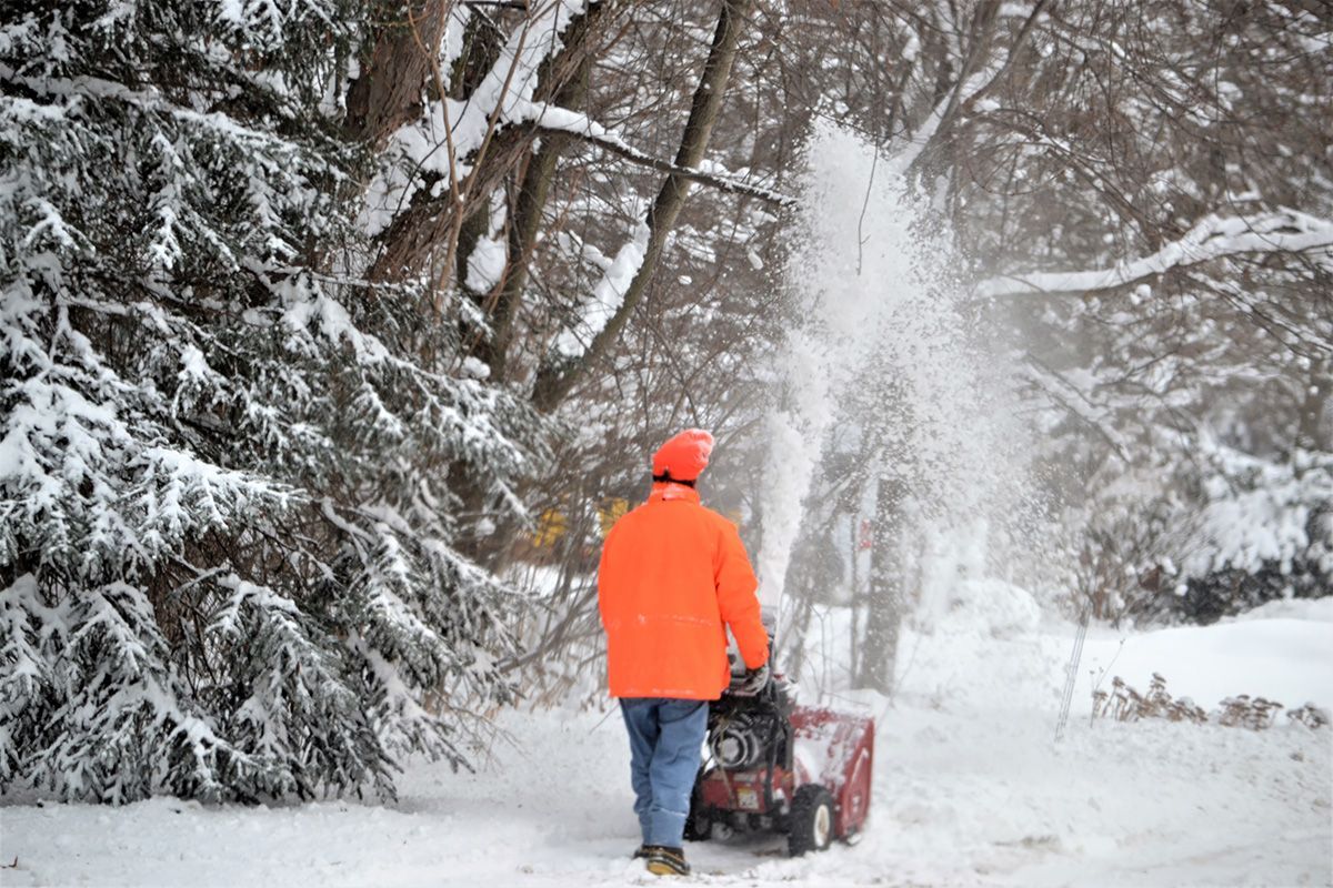 A man in an orange jacket is blowing snow from a snow blower.