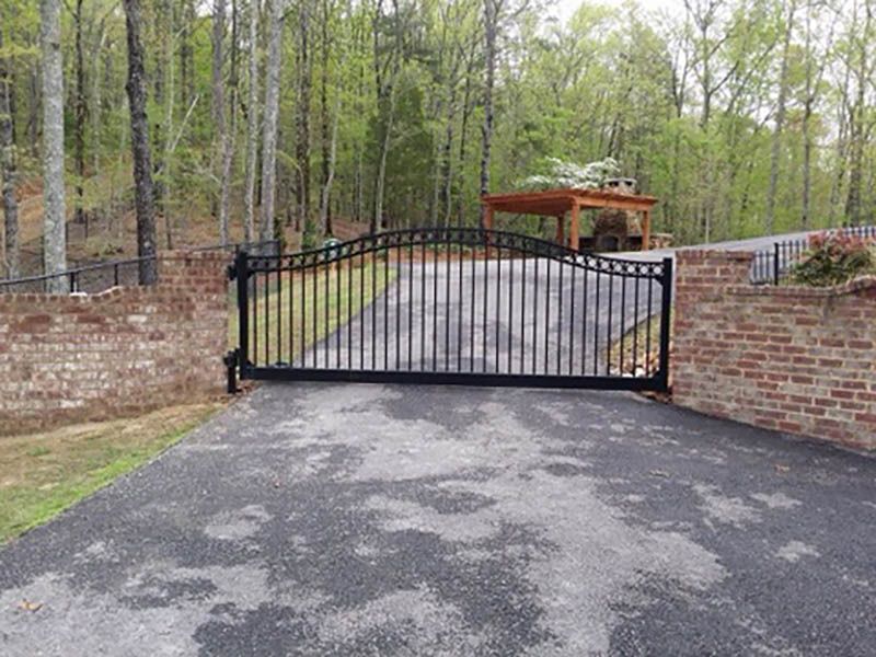 A black metal gate is open to a driveway with trees in the background.