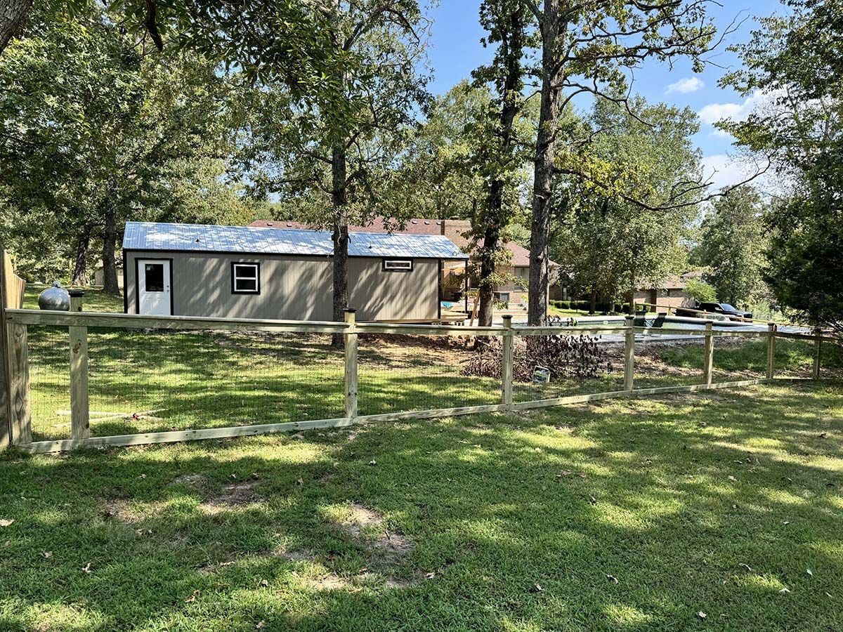 A small house with a wooden fence in front of it is surrounded by trees.