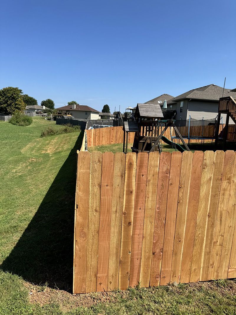 A wooden fence surrounds a backyard with a playground in the background.