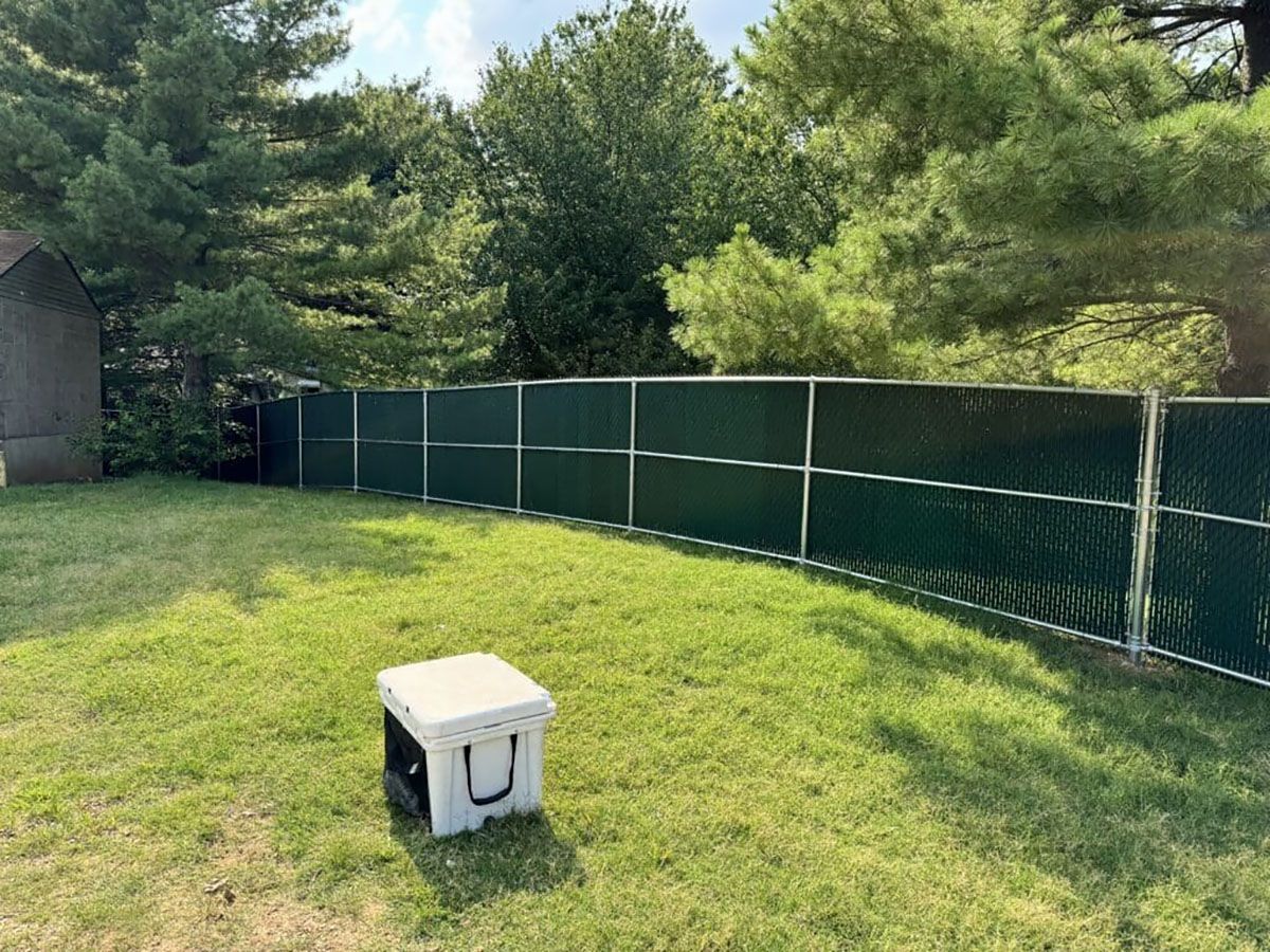 A white cooler is sitting in the grass in front of a green fence.