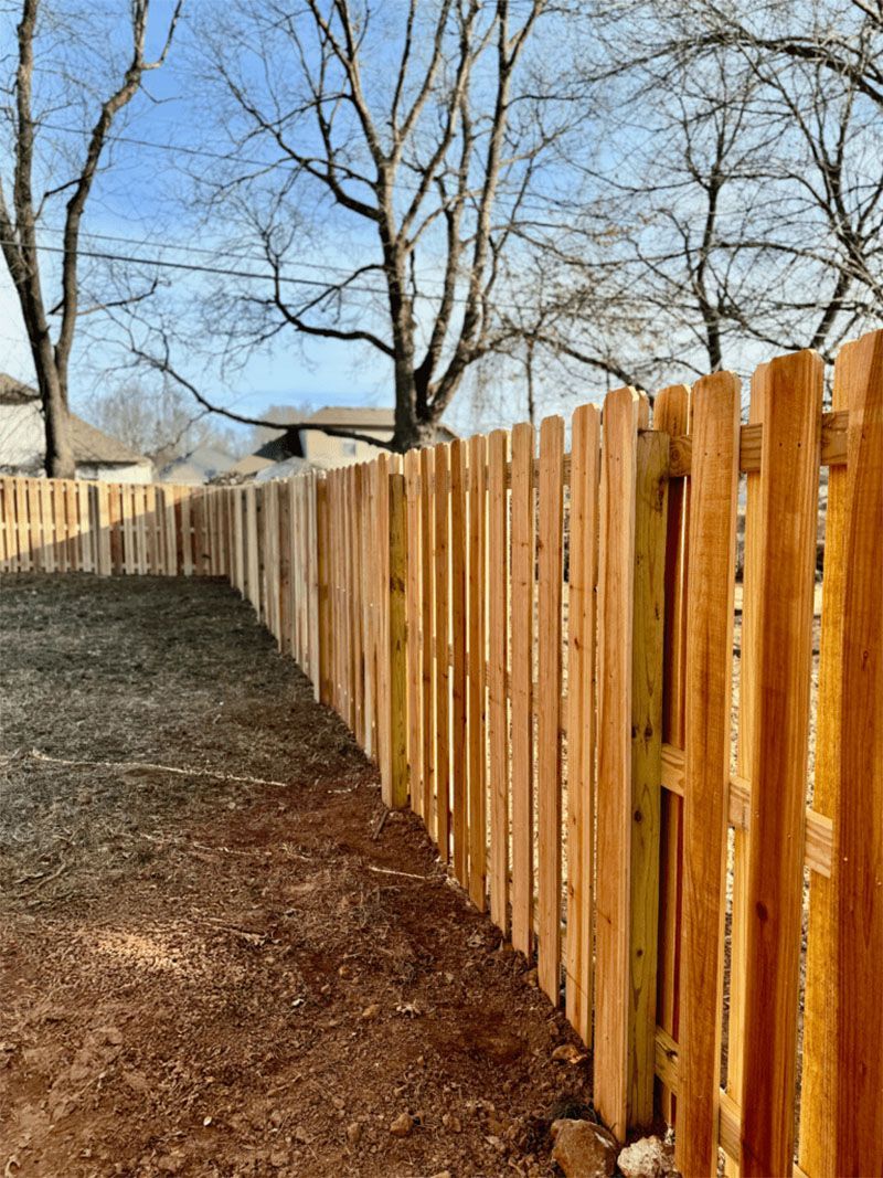 A wooden picket fence surrounds a dirt yard with trees in the background.