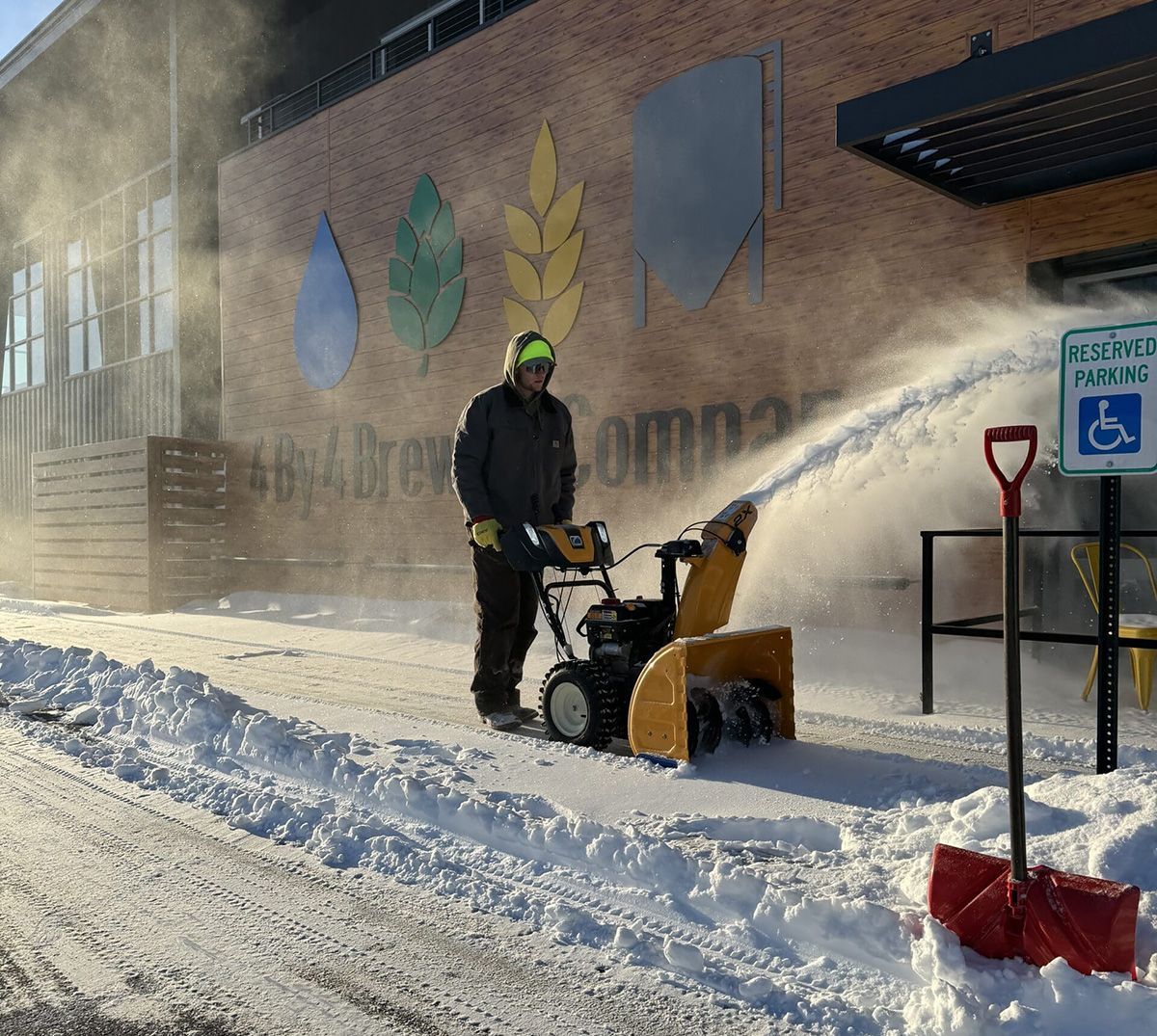 A man is blowing snow from a snow blower in front of a handicapped parking sign