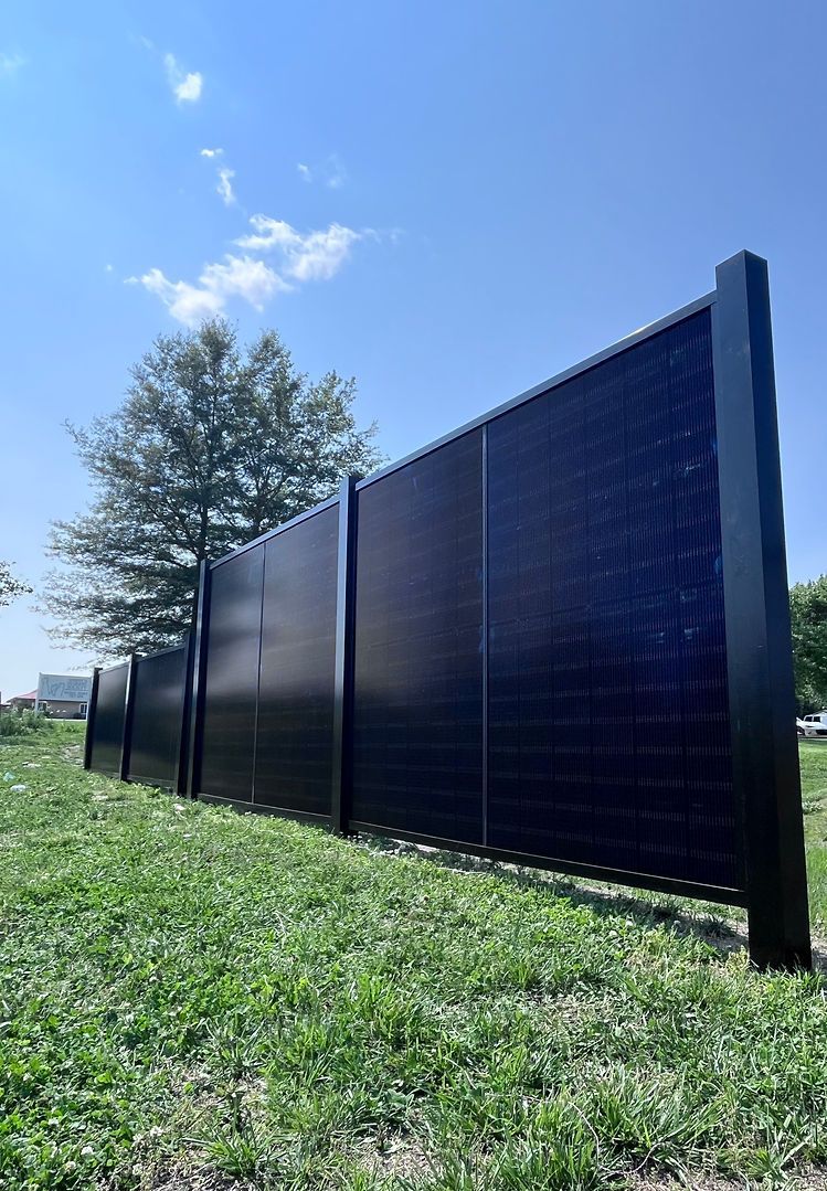 A black fence with solar panels on it is sitting in the middle of a grassy field.