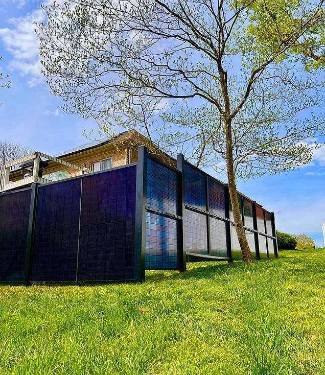 A black fence with solar panels on it is in the grass in front of a house.