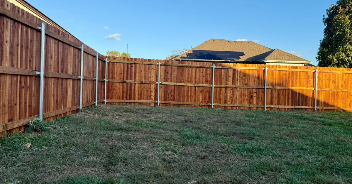 Brown wooden fence encloses a grassy backyard under a clear blue sky. A house is in the background.