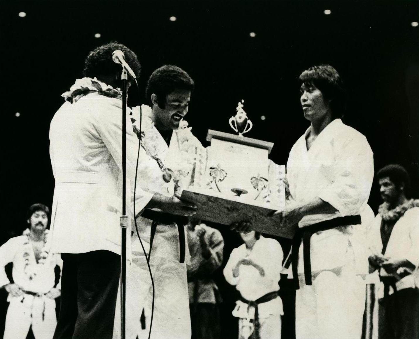 Karate tournament award ceremony. Two men in white gis hold up a trophy on a stage.