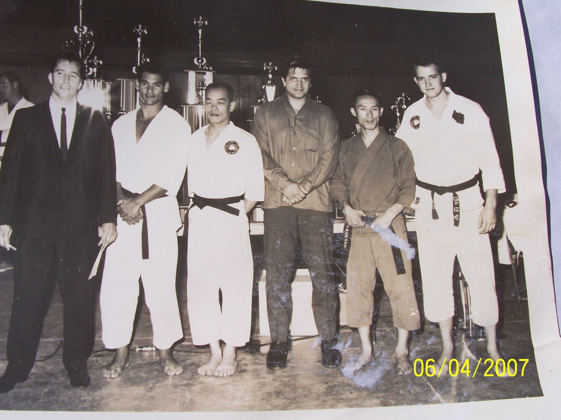 Group of six men, some in karate uniforms, standing together indoors with trophies in the background.