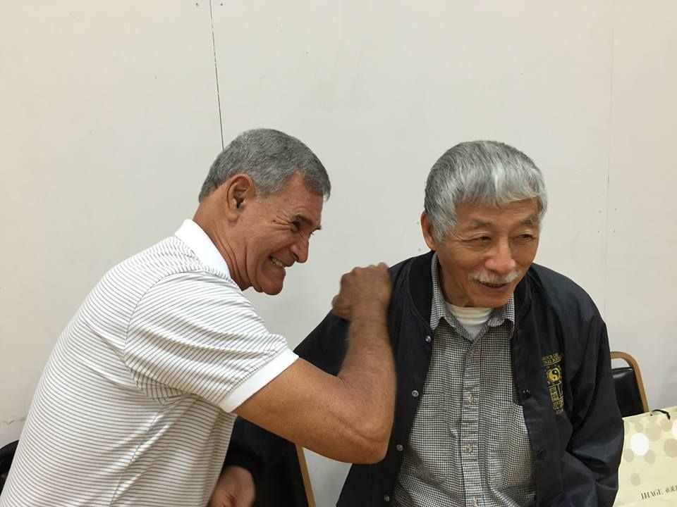 Two men, one smiling with arm around the other's shoulder. They are in front of a plain wall.