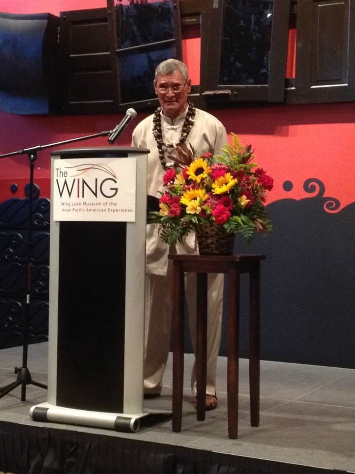 Man in a beige suit stands at a podium with flowers, speaking at an event labeled