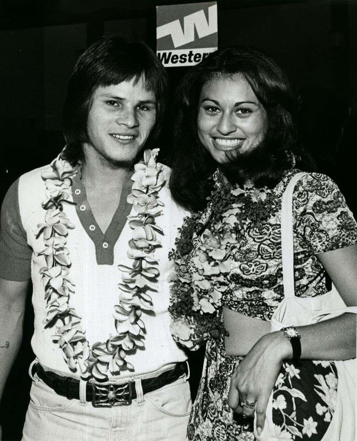 A man and woman wearing leis pose in front of a Western Airlines sign.