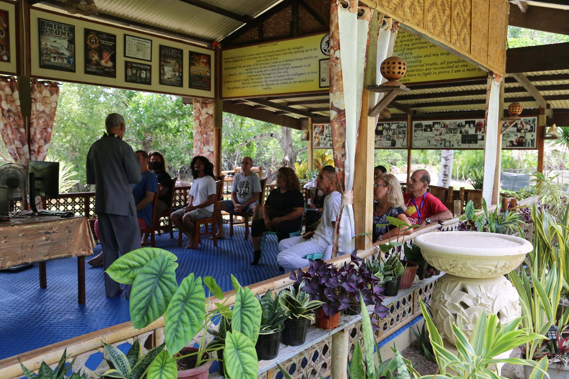 A group of people sit and listen to a speaker under an outdoor pavilion with plants.