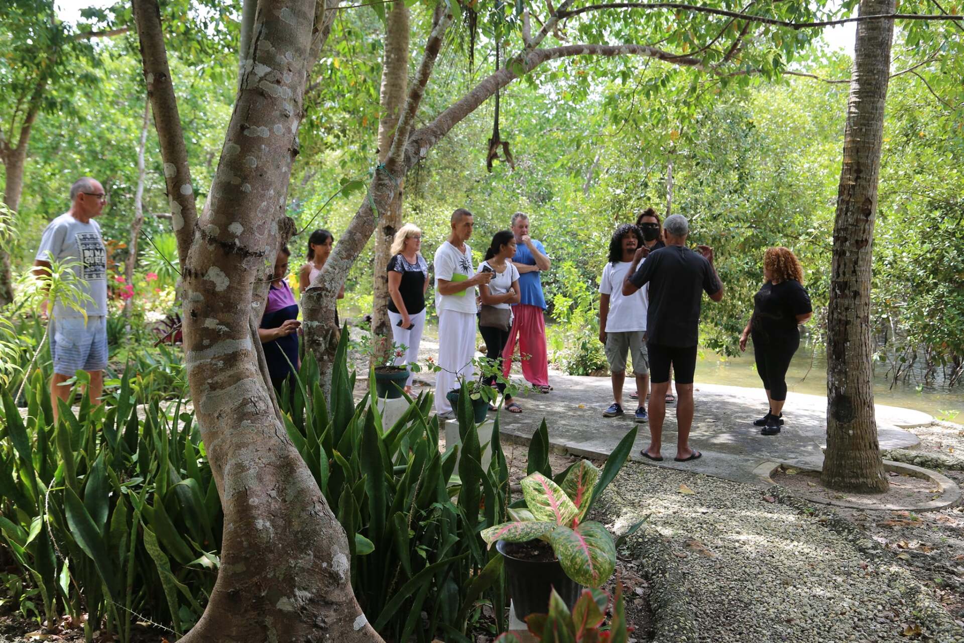 A group of people gathered outdoors, listening to a man in white clothing speaking. Lush greenery surrounds them.