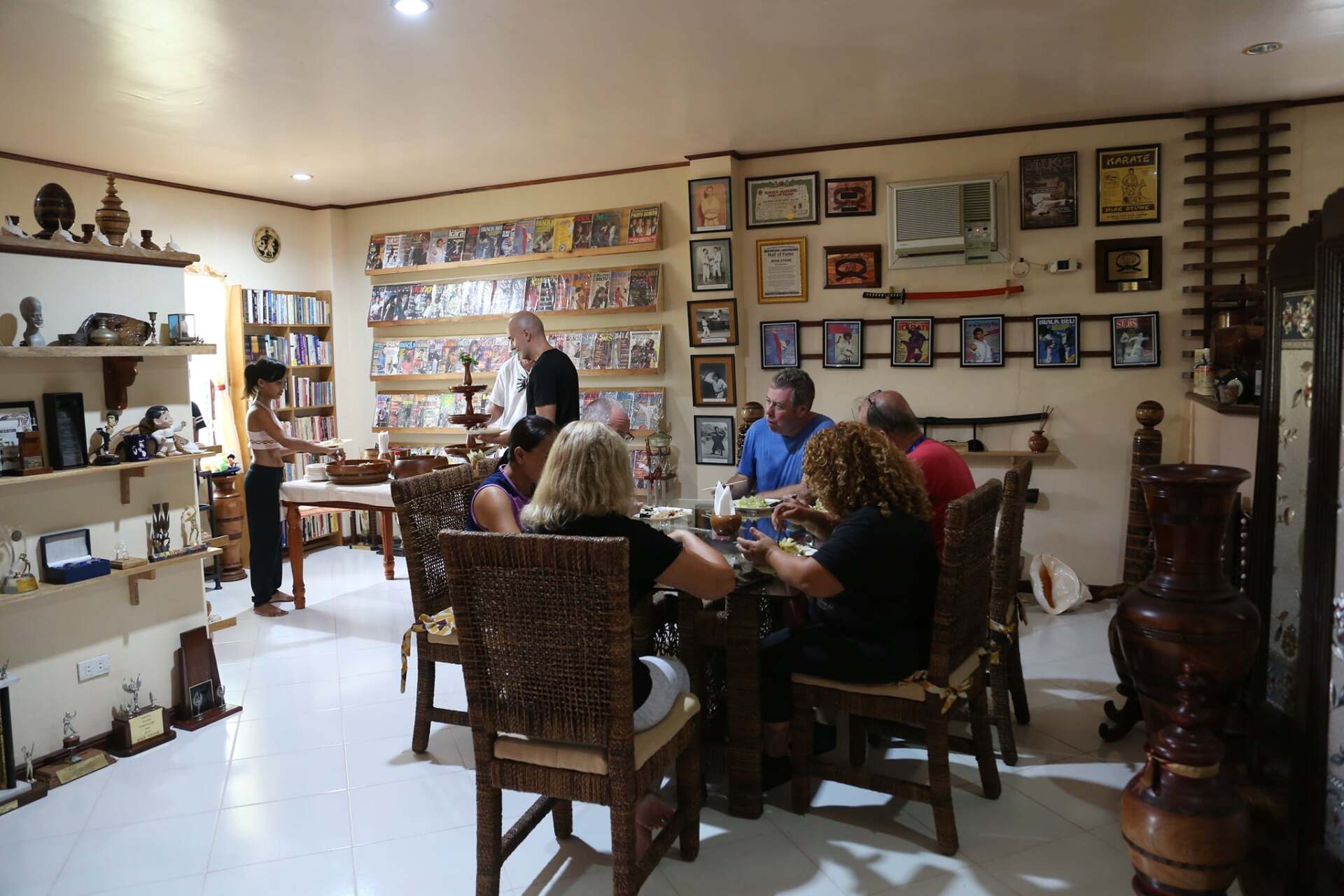 A group of people seated around a wooden table in a dining room, eating. Walls are decorated with framed photos and artwork.