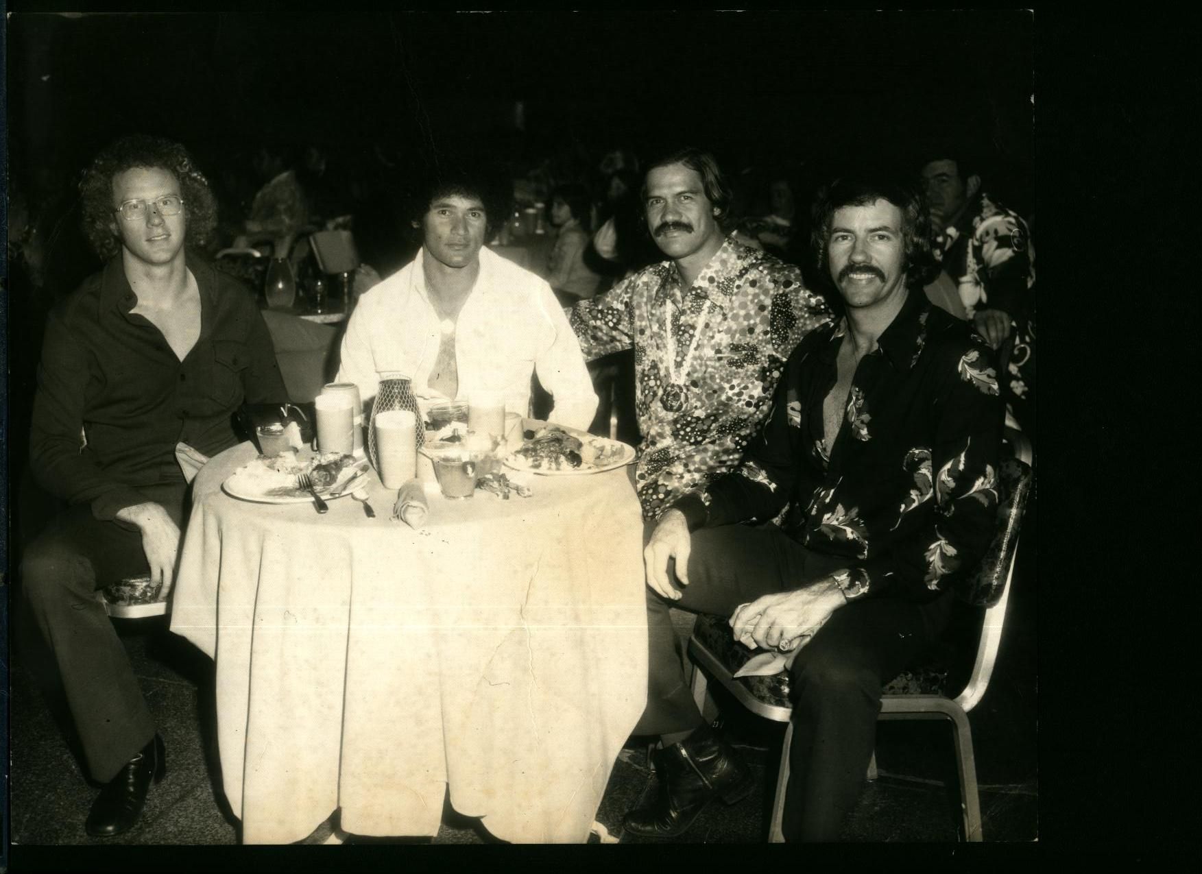Four men seated at a round table in a dimly lit room. They are smiling and wearing casual attire.