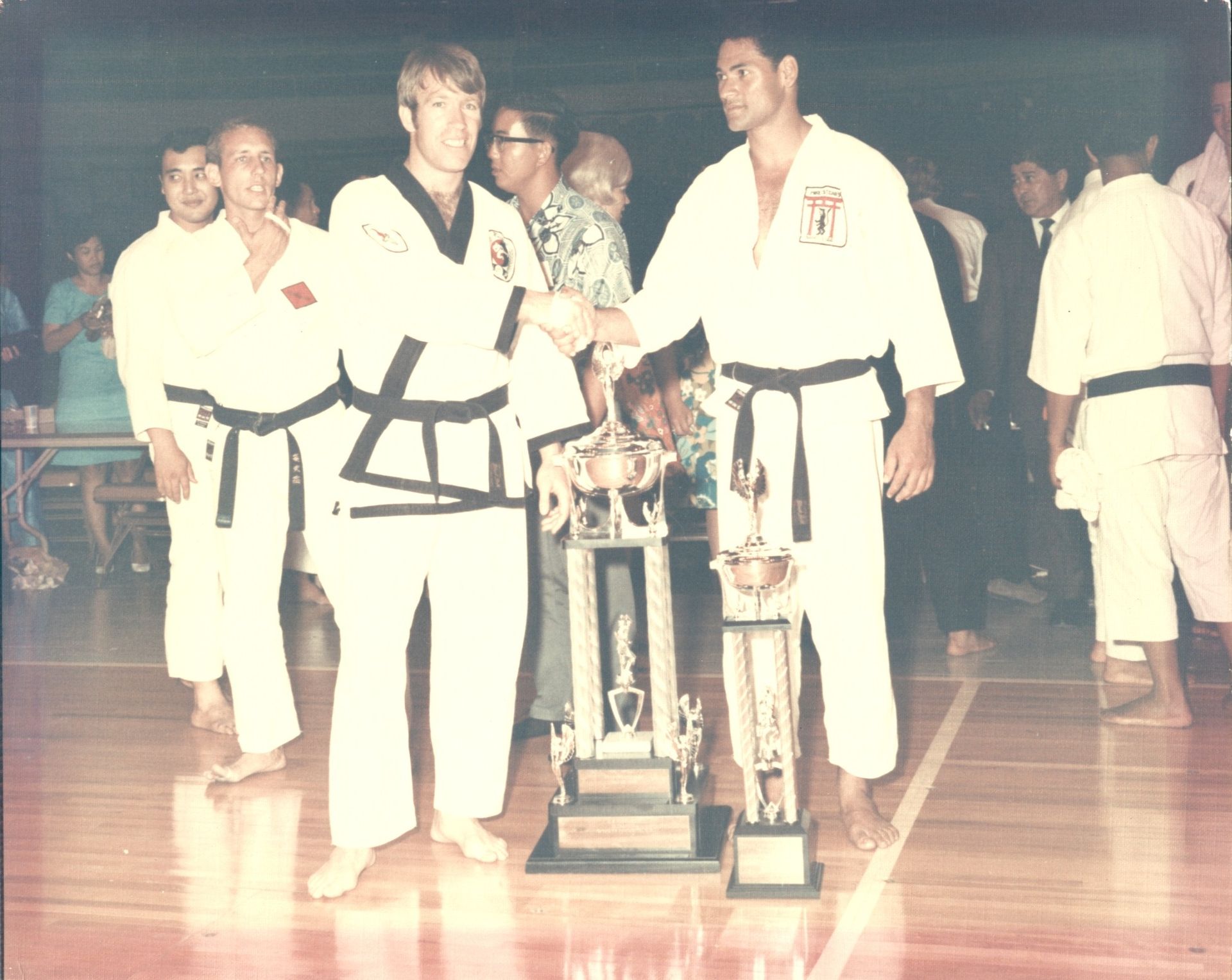 Two martial artists in white uniforms and black belts shake hands, holding trophies at a competition, likely indoors.