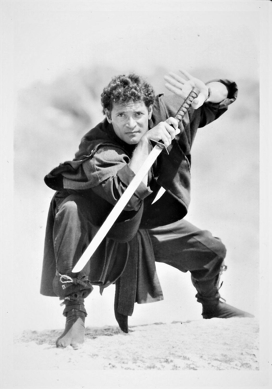 Man in ninja attire, posing with a katana in an outdoor setting. He holds the sword with both hands, ready to strike.