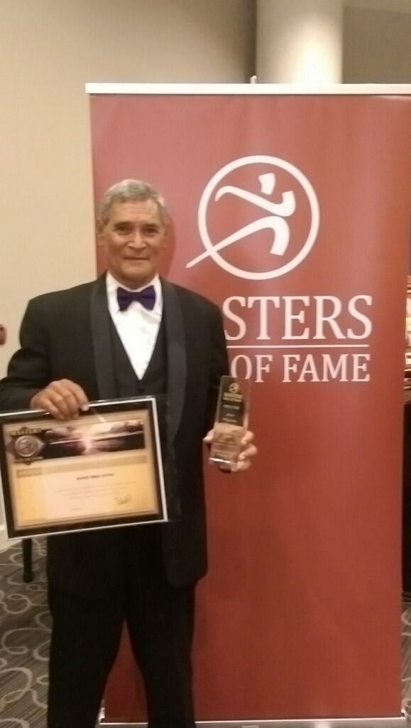 Man in a tuxedo holds an award and plaque in front of a 