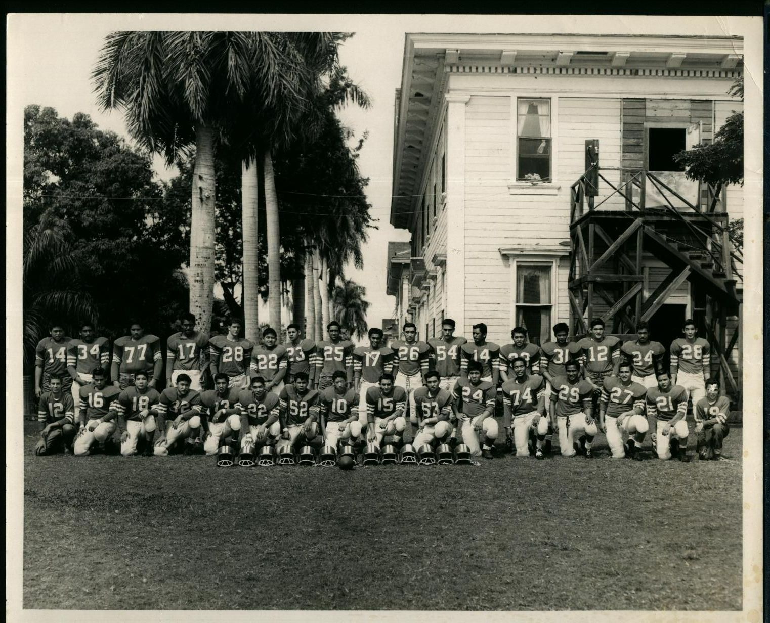 Football team posing for a team photo in front of a large building and tall trees. Players are wearing uniforms.