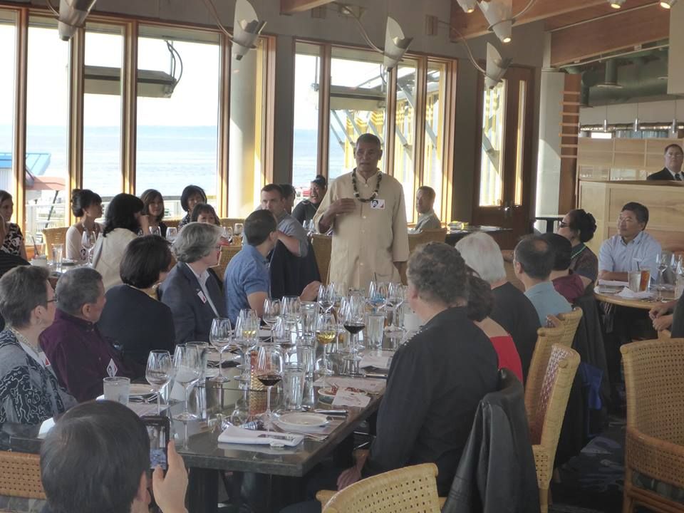 A man addresses a group at a restaurant table, holding glasses, gesturing.