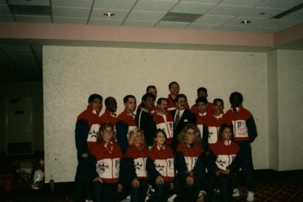 Group of people in red, white, and blue jackets posing with a man in a suit in front of a plain wall.