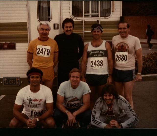 Group of seven men posing in front of a camper, likely before a race. They wear running attire, with bib numbers visible.