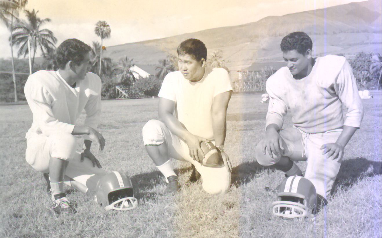 Three football players in vintage uniforms on a grassy field, kneeling with helmets and a football.