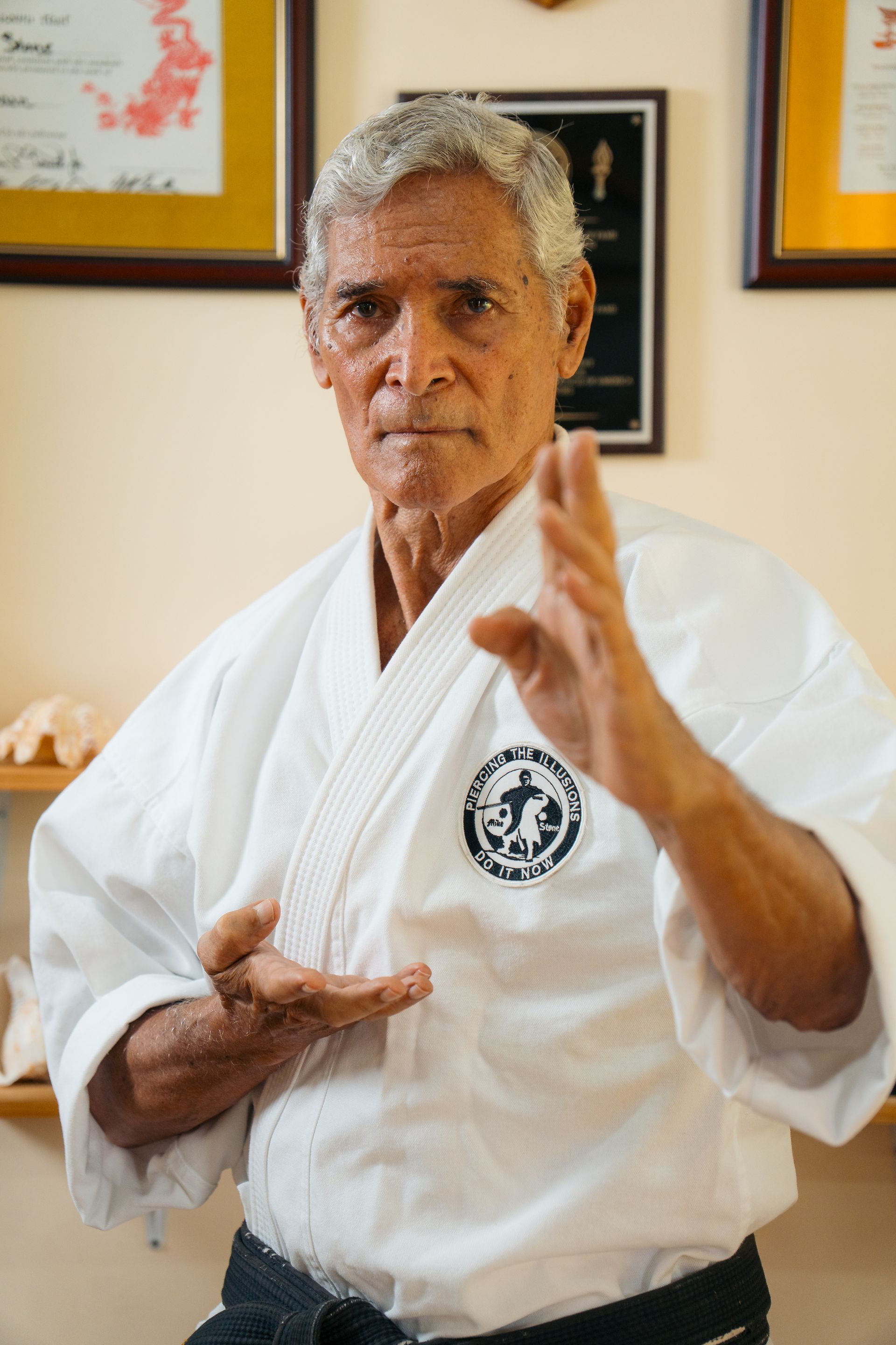 Elderly man in karate uniform, hands up in defensive posture, in a dojo with certificates on the wall.