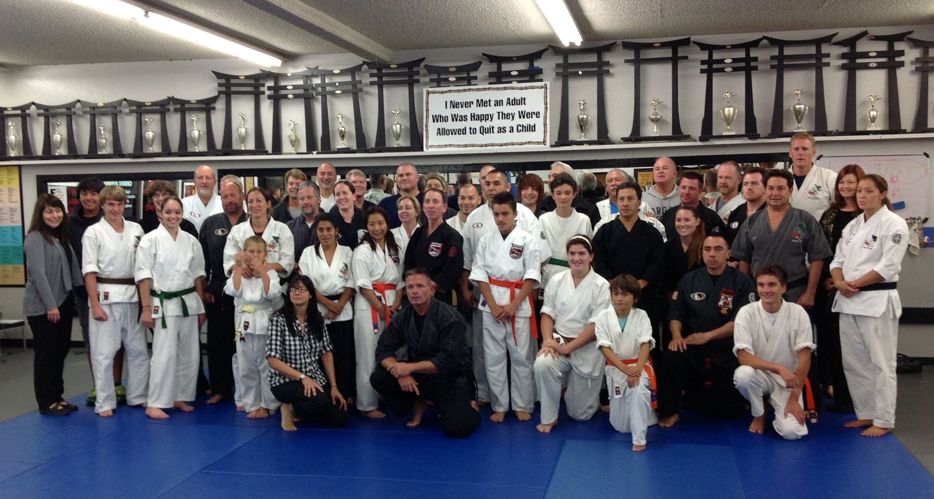 Group of martial arts practitioners in a dojo, many wearing white uniforms with belts; some in black. 