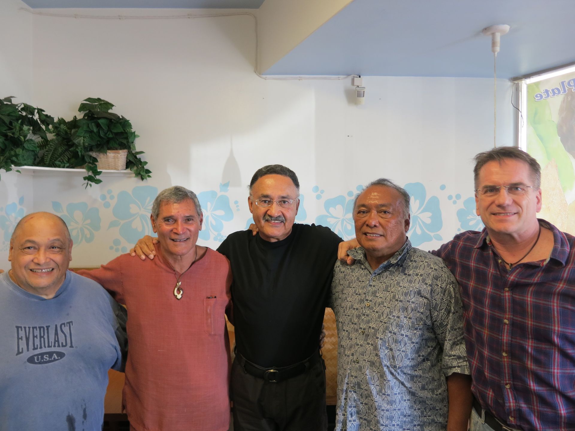 Five men stand close together, smiling, with arms around each other. They are in a room with a plant and a map on the wall.