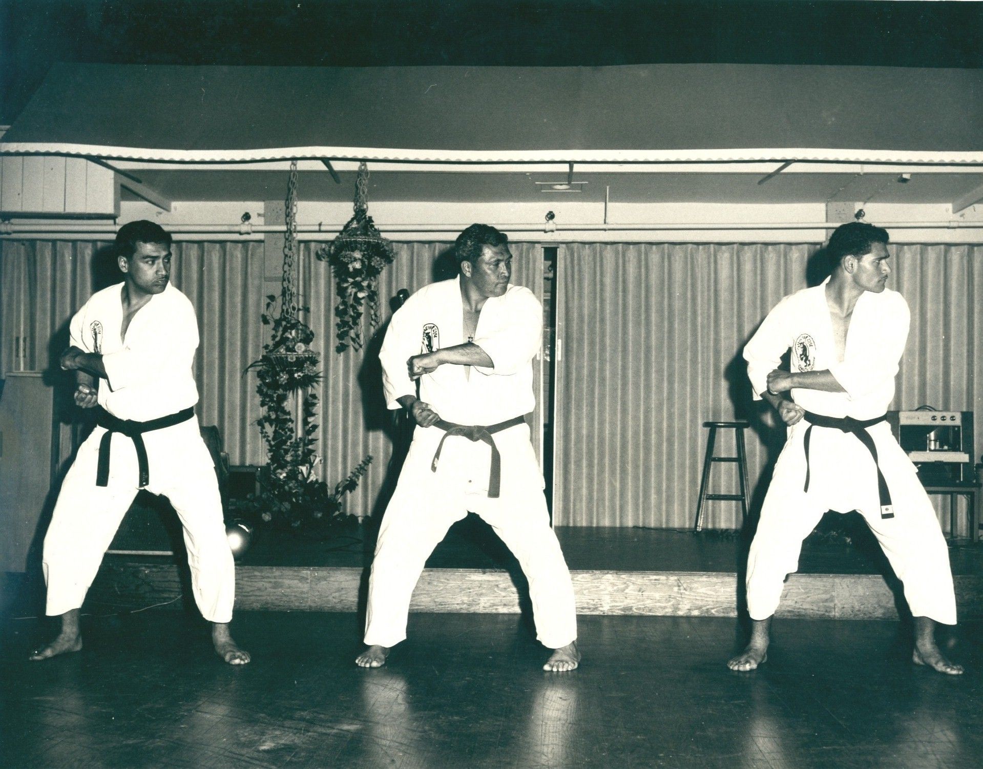 Three people in white karate gis with black and brown belts practicing a kata indoors.
