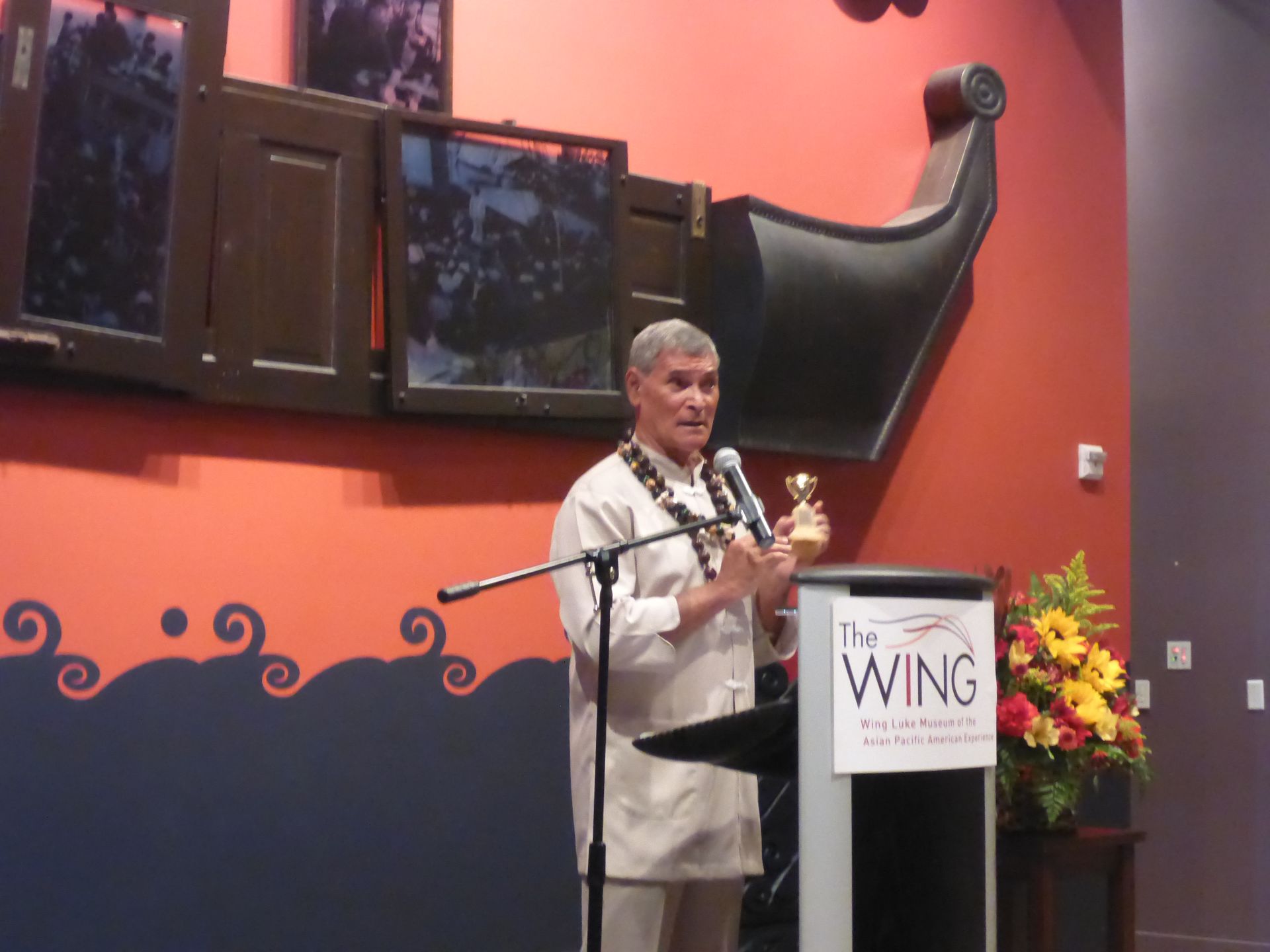 Man at a podium holding a small trophy, speaking at The Wing Luke Museum. 