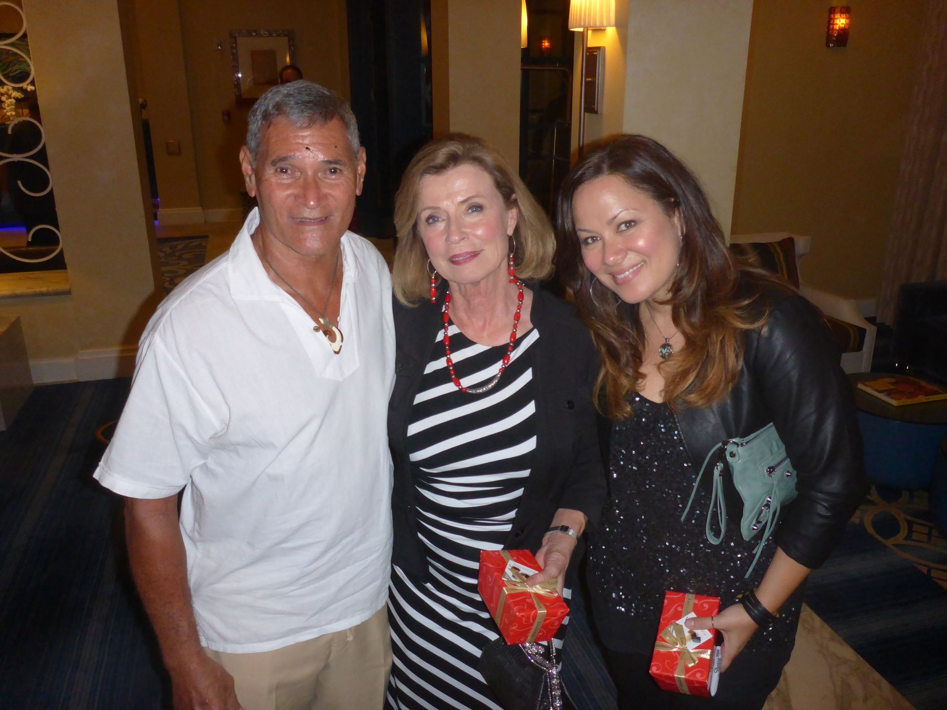 Three people posing for a photo indoors: a man in white. They are holding red gift boxes and smiling.