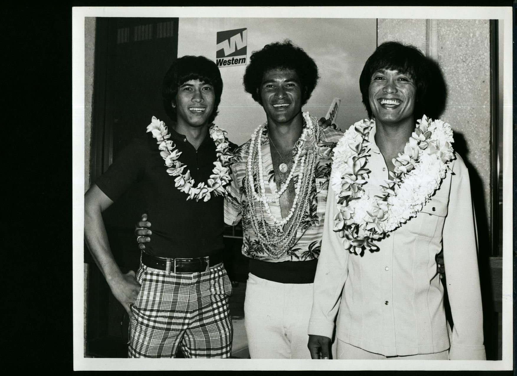 Three men in leis pose for a photo. They have dark hair and are dressed in casual attire.