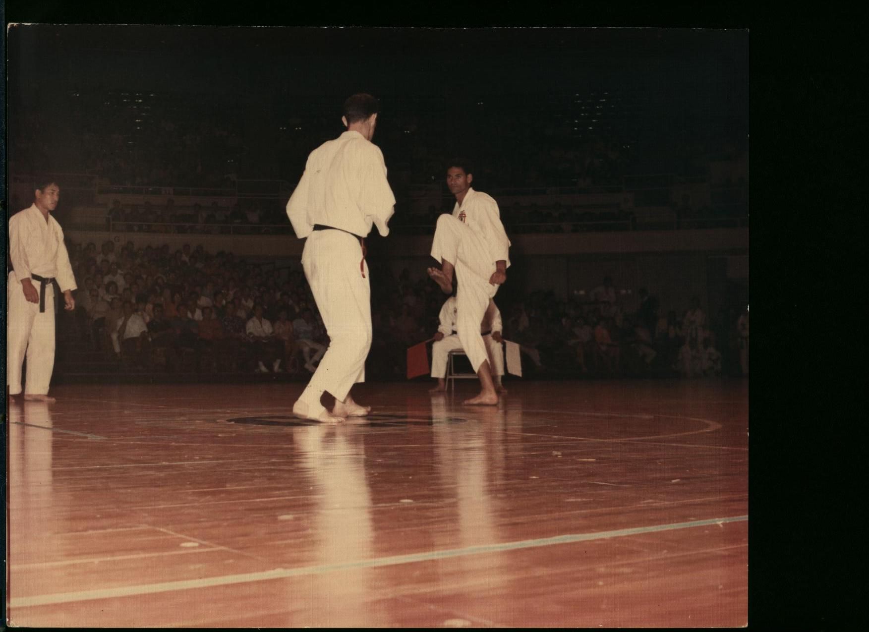 Two karate practitioners sparring on a wooden floor in a large indoor arena.