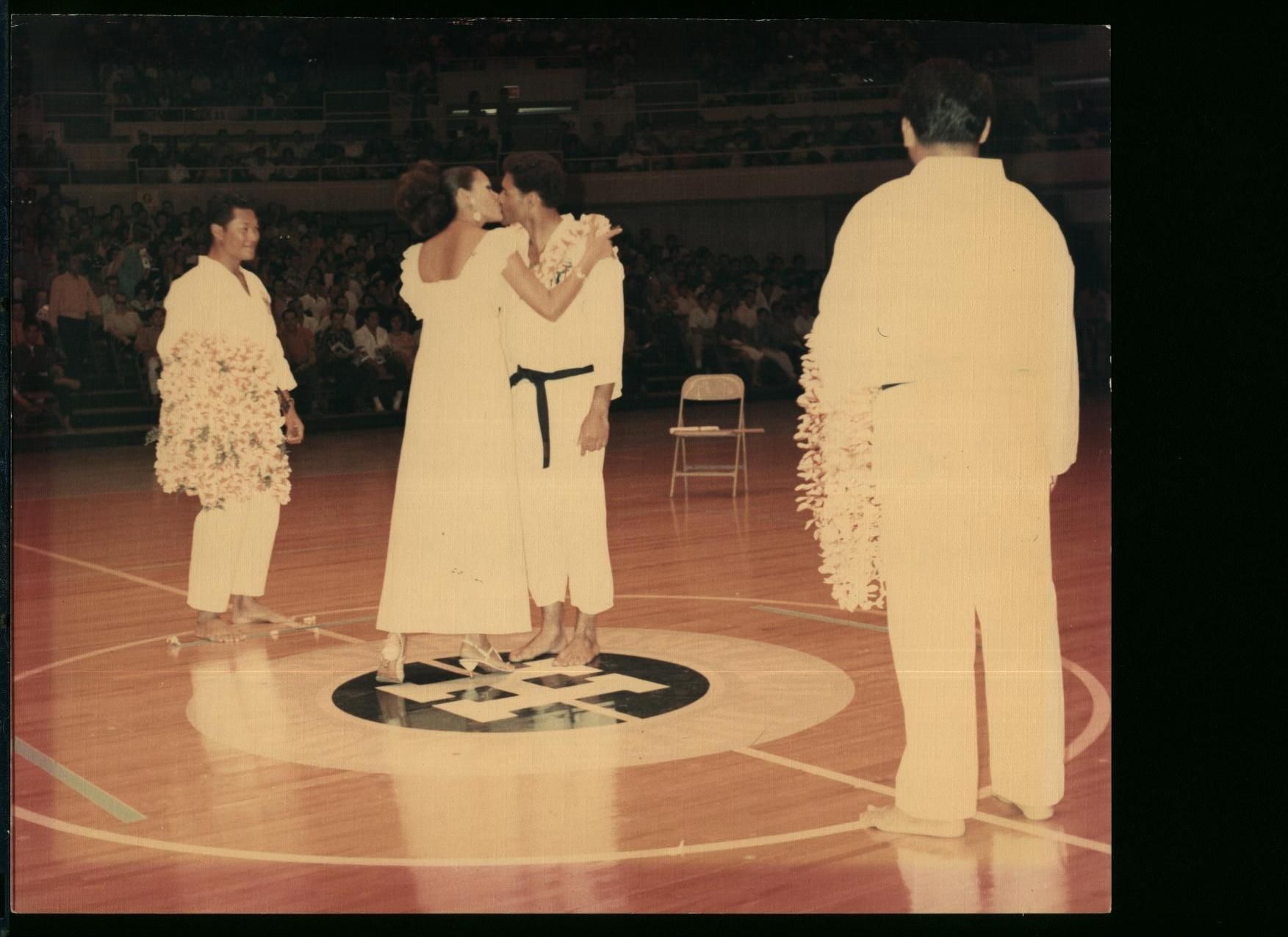 Two people in karate uniforms embracing on a basketball court, while others in white karate attire watch;