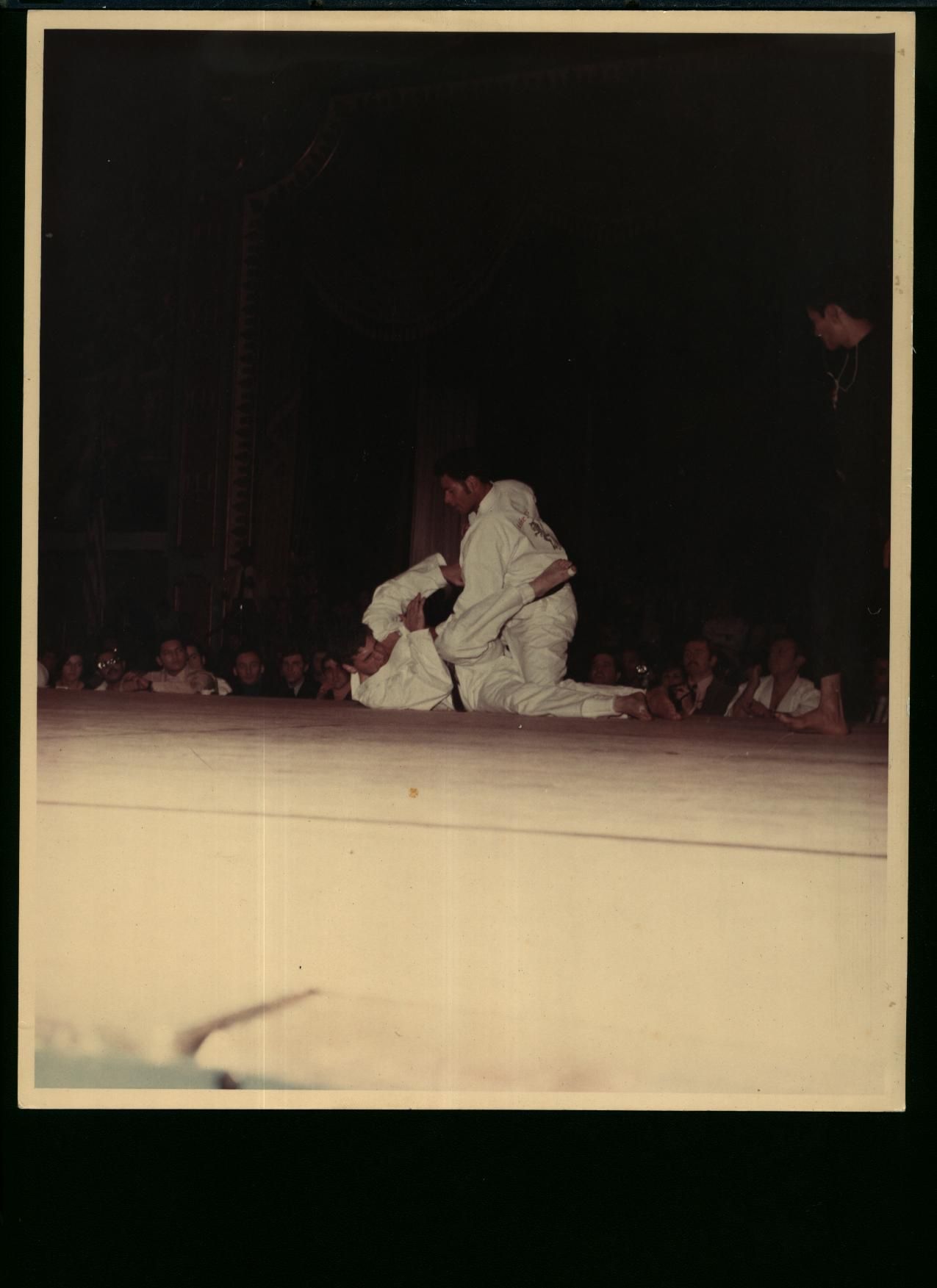 Two people in white judo uniforms grapple on a stage, watched by a crowd. Dim lighting, dark background.