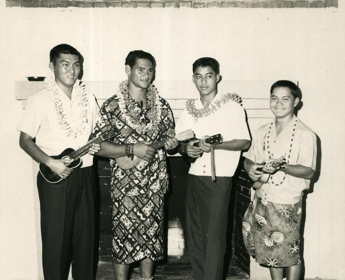 Four men in traditional attire holding ukuleles, possibly a band, in front of a fireplace. They wear leis and are smiling.