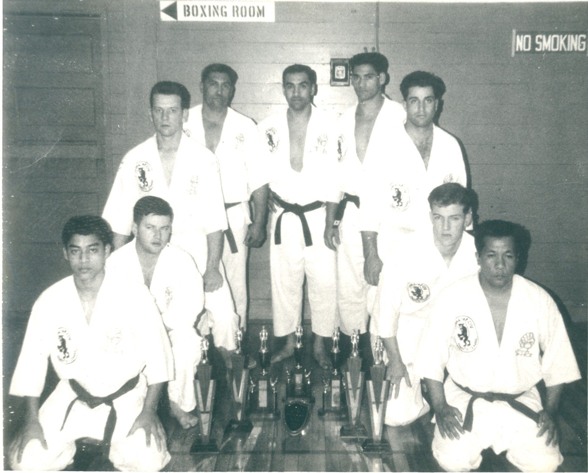 Group of karate practitioners in white uniforms pose with trophies in a gymnasium. A