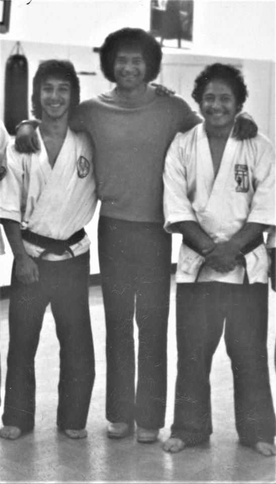 Three men posing. Two on the ends wear karate gis, one in the middle in casual clothes, all smiling.