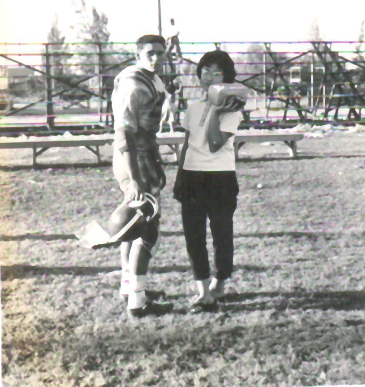 A man in football gear and a woman stand together on a grass field. The woman carries a box on her shoulder.