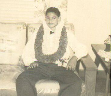 A young man wearing a lei and a bow tie sits on a couch, smiling. The photo is in black and white.