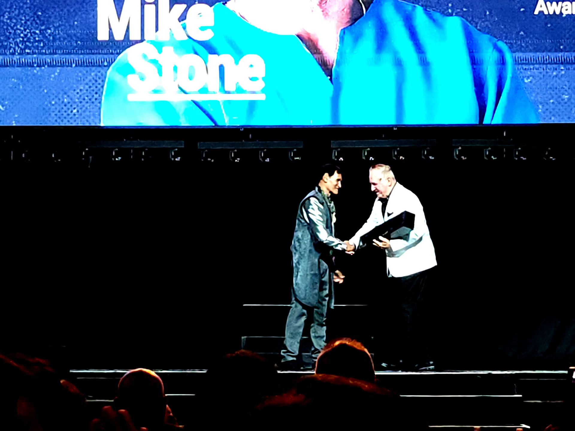 A man in a blue jumpsuit shakes hands with another man on a stage under a screen that says 