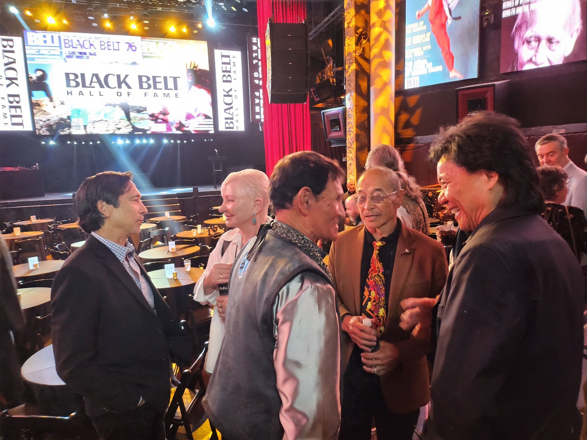 A group of people conversing indoors, potentially at an event with a stage visible in the background. 