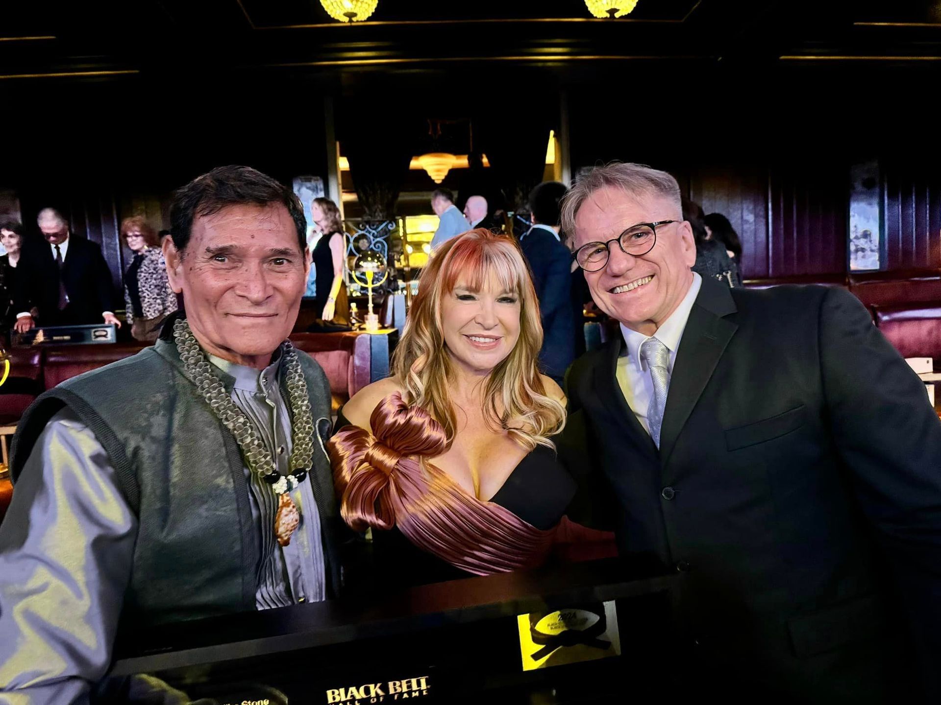 Three people pose at an event: a man in an ornate outfit, a woman in a gown, and a man in a suit, smiling. They are indoors.