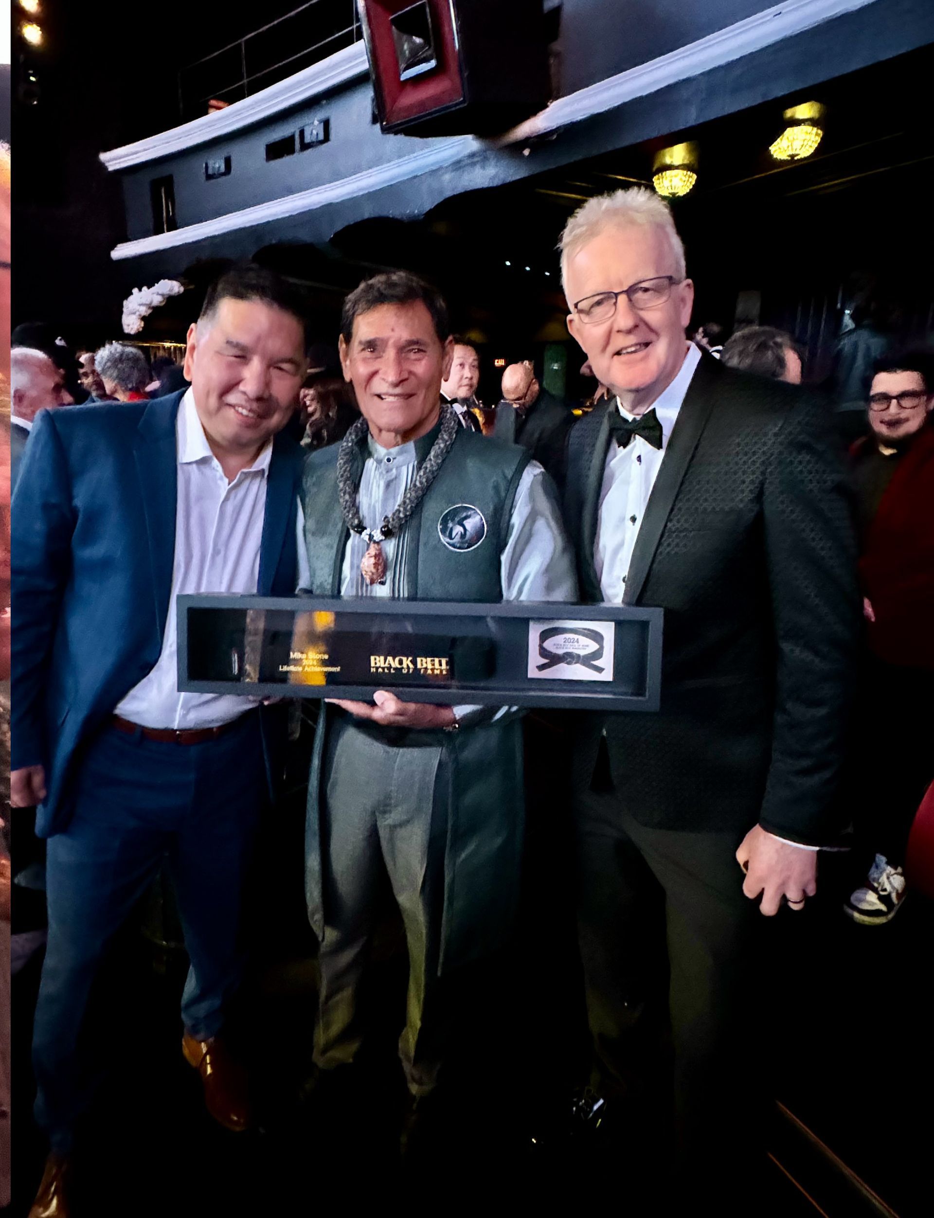 Three men in formal attire holding an award. The man in the middle is smiling, holding the encased award; others smile.