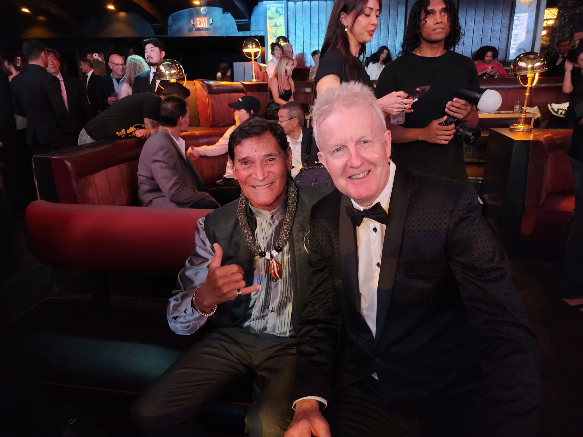 Two men smiling, seated on a plush bench at a dimly lit event. One gives the shaka sign, the other in a tuxedo.