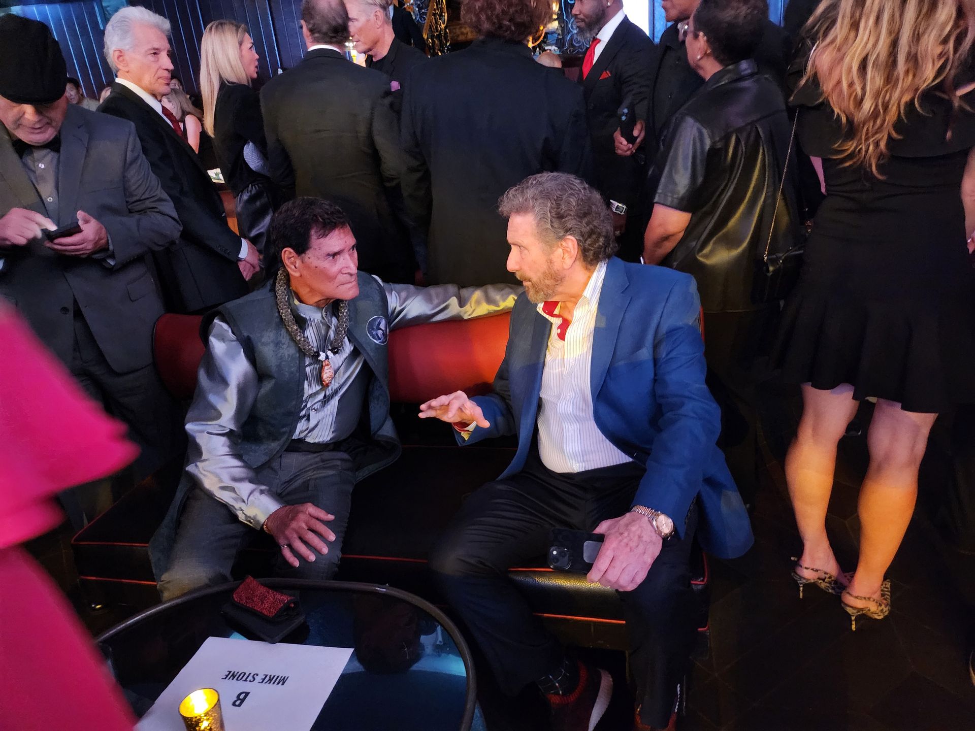 Two men seated on a red velvet couch in a crowded setting, one in a blue suit and the other in a patterned vest.