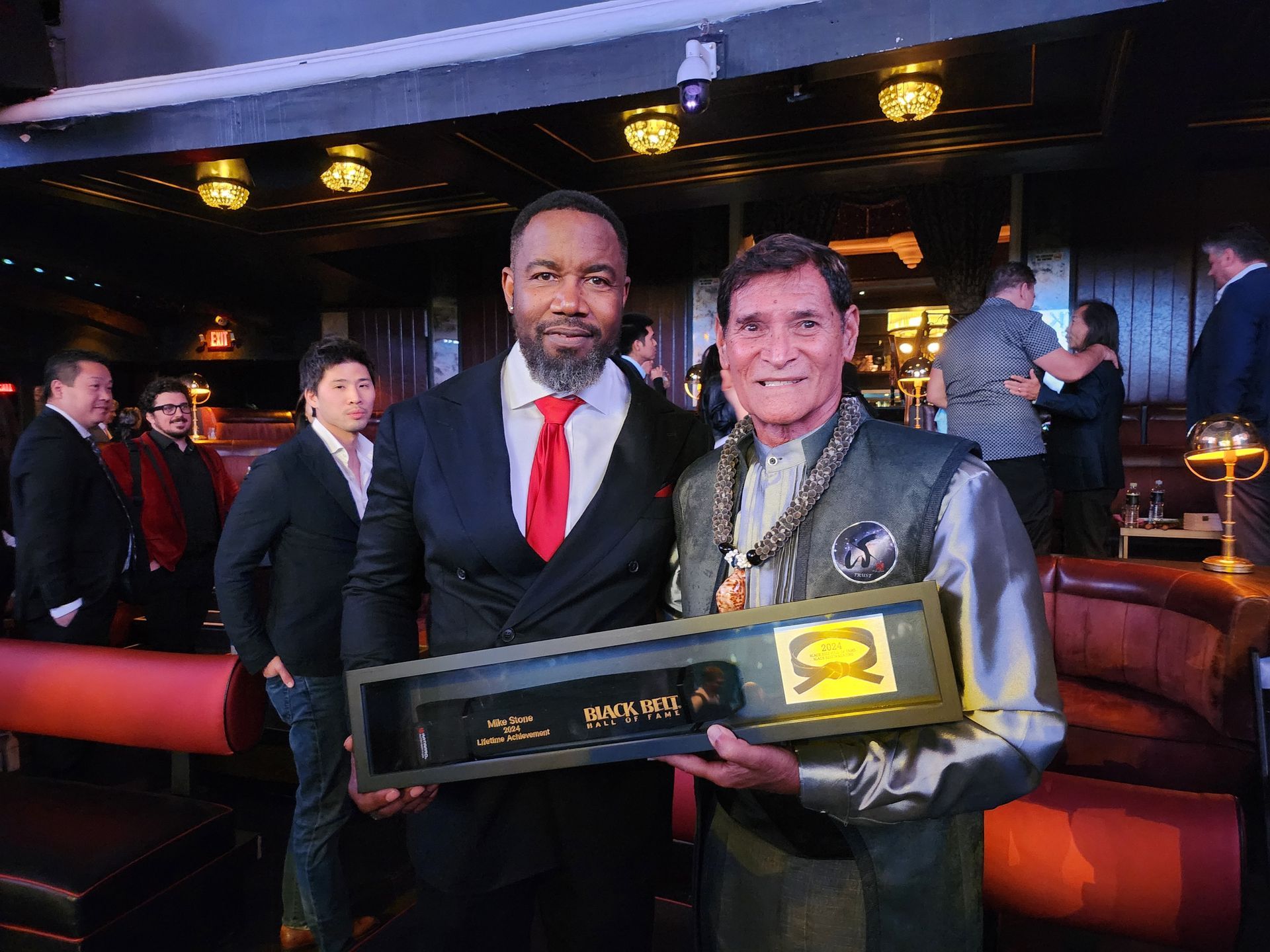Two men indoors pose with a framed award. One man wears a suit, the other a decorated top. Dimly lit setting.
